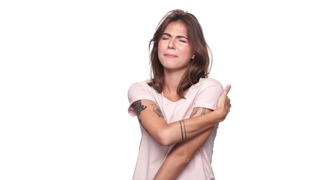 Confused brunette woman in t-shirt having pain in shoulder over white background
