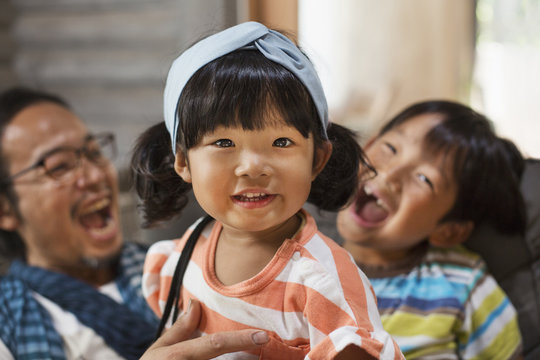 Man, boy and young girl with black pigtails wearing blue hairband sitting on a grey sofa, laughing.