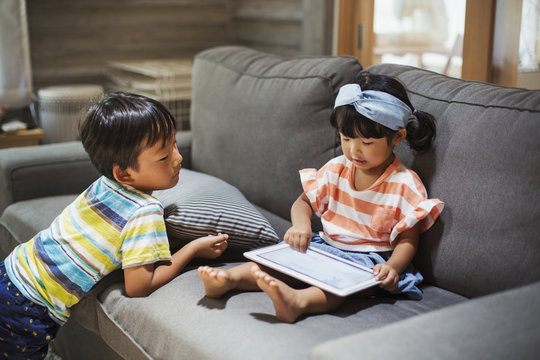 Young girl with black pigtails sitting on a sofa, digital tablet on her lap, boy standing beside her.