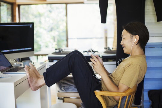 Side view of man sitting indoors at a desk with computer, legs raised, barefoot, holding mobile phone, wearing headphones.
