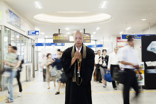 Buddhist monk standing inside train station, holding mala, people walking past