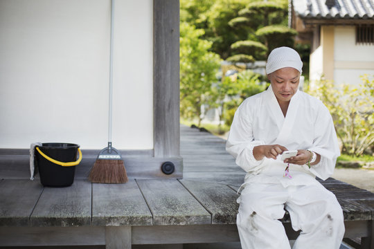Buddhist monk wearing white robe and cap sitting on wooden floor outside a temple, using mobile phone.