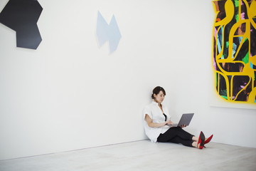 Woman with short black hair wearing white shirt, black trousers and red high heel shoes sitting on floor i art gallery, holding laptop.