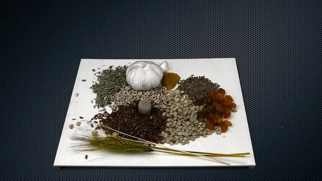Nuts fall on grains on background of metallic dark gray grid. Eco-friendly foods for health. Seeds wheat lentils cumin raisins garlic and other products on wooden board. Slow motion.