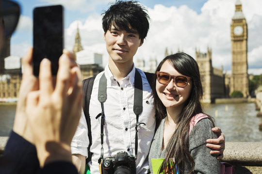 Smiling woman with black hair taking picture of couple with smartphone, standing on Westminster Bridge over the River Thames, London, with the Houses of Parliament and Big Ben in the background.