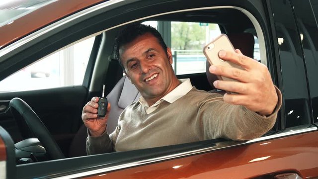 A man is sitting at the wheel of a car. He takes out the phone and the keys to the new car. Then he makes a selfie. He smiles very broadly