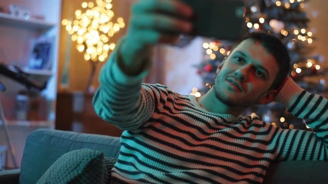Young man in striped sweater sitting on couch in living room on background of nice Christmas tree. Man taking selfie with holiday gift, using smartphone camera.