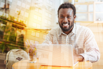 Be positive. Handsome dark-skinned man sitting at his workplace and looking at camera, using his laptop