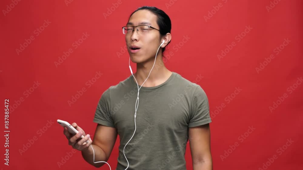 Sticker Pleased asian man in t-shirt and eyeglasses holding smartphone and listening music from headphones over red background