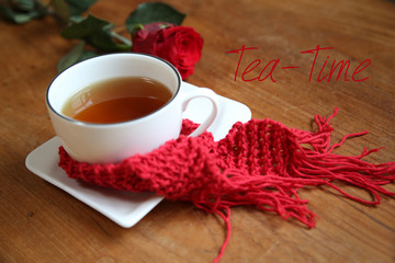 Eine tsse mit Tee umwicklt von einem roten Schal und einer Rose nebenlieden und dem Text "Tea-Time"