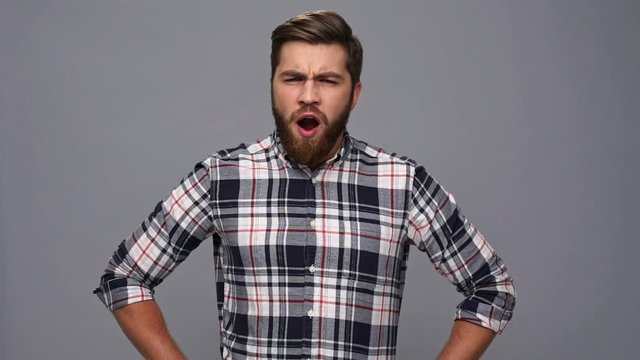 Tired bearded man in shirt yawns and holding arms on hips over gray background
