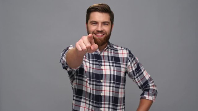 Happy bearded man in shirt holding arms on hips and pointing at the camera over gray background
