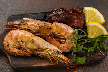 Fried tiger shrimps with lemon, sun-dried tomatoes and greens or rukkola on a dark wooden cutting board.