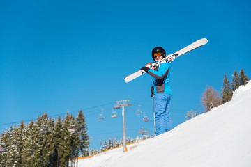 Low angle shot of female skier on the hill in snowy mountains holding her skis on the shoulder. Blue sky, winter forest and ski resort on the background copyspace lifestyle sportswoman weekend concept