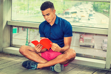 Way to Success. I Love Reading Book. African American college student sitting cross legged on floor against glass wall on campus, looking down, reading red book. Street, many cars on background..