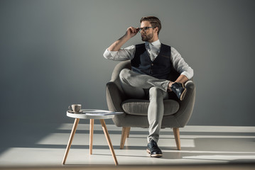 full length view of handsome stylish young man sitting in armchair and looking away