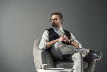 portrait of handsome young businessman in eyeglasses looking away while sitting in armchair isolated on grey