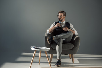 handsome stylish young man sitting in armchair and looking away while drinking coffee