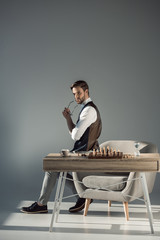 pensive stylish young businessman holding eyeglasses and looking away while playing chess