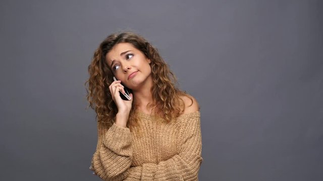 Upset curly woman in sweater talking by smartphone over purple background