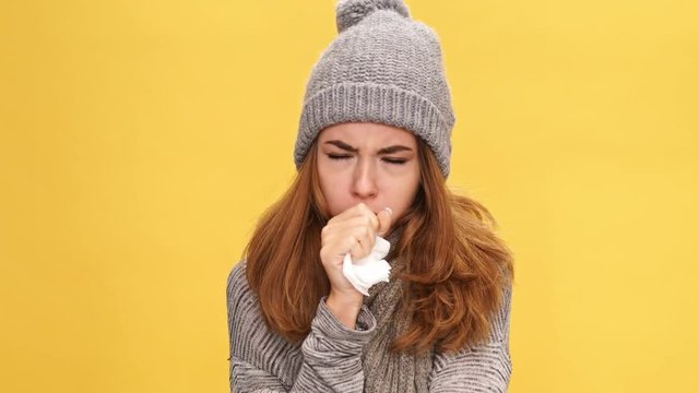 Confused brunette woman in sweater and hat having runny nose and coughs over yellow background
