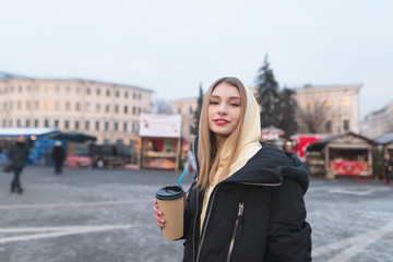Naklejka na ściany i meble A positive girl with a glass of coffee in her hands stands on the background of the fair, smiles and looks at the camera.