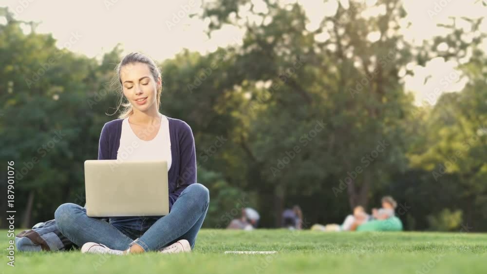 Poster pleased brunette woman sitting on grass and using laptop computer in park