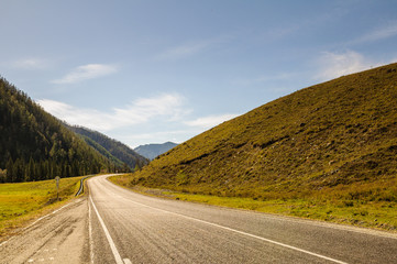 Road through a field of orange grass on highland steppe on a background of snow covered high mountains and glaciers under clouds and blue sky, Altai mountains, Siberia