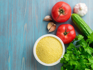 Vegetarian food - wheat, grain of raw couscous in a white bowl on a blue wooden background, fresh vegetables - parsley, cucumbers, tomatoes, top view
