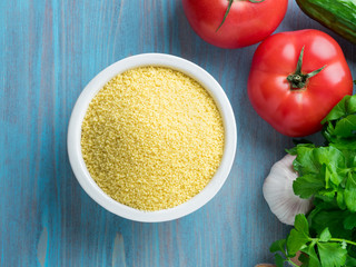 Vegetarian food - wheat, grain of raw couscous in a white bowl on a blue wooden background, fresh vegetables - parsley, cucumbers, tomatoes, top view