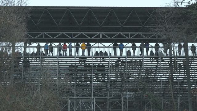 Group of spectators standing on the bleachers, bare trees in foreground.
