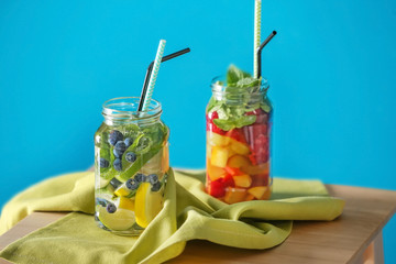Mason jars of infused water with fruits and berries on wooden table against color background