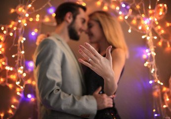 Young man and his happy fiancee showing engagement ring against blurred lights