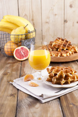 Sweet Belgian waffles and a glass of orange juice for breakfast and bananas in a basket, on a wooden plate. Homemade baking. On a brown wooden background. Free space for text or advertising