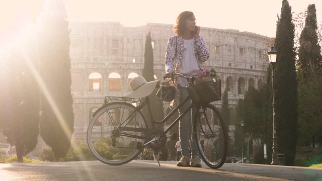 Beautiful young woman in colorful fashion dress with bike talking on phone call in front of colosseum in Rome at sunset with trees happy attractive tourist girl in colle oppio