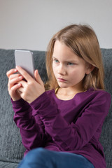 Mädchen schaut auf ein Smartphone und sitzt auf einem grauen Sofa