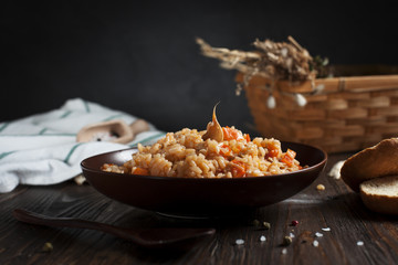 pilau in a brown ceramic dish, a kitchen towel, cherry tomatoes and a basket with spikelets of wheat, bread and a wooden spoon on a dark wooden table