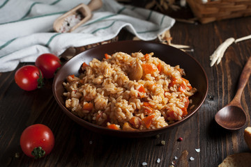 pilau in a brown ceramic dish, a kitchen towel, cherry tomatoes and a basket with spikelets of wheat, bread and a wooden spoon on a dark wooden table