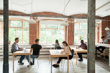 Multiracial people work in modern co-working center interior, diverse employees collaborate sitting at desks of loft shared office, african caucasian colleagues talking in coworking space, teamwork