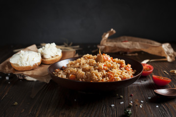 pilaf in a brown ceramic plate, cherry tomatoes, toast with feta cheese and a wooden spoon on a dark wooden table