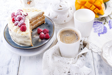 Cut the cake with white cream, for breakfast. A mango fruit. White background, tablecloth with lace, a cup of fragrant black coffee and free space for text or advertising.