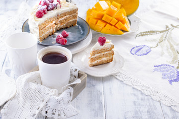 Cut the cake with white cream, for breakfast. A mango fruit. White background, tablecloth with lace, a cup of coffee and free space for text or advertising.