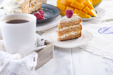 Portion of cake on a small white plate, with a cream for breakfast. A mango fruit. White background, cup of coffee and free space for text or advertising.