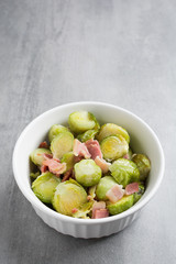 Brussel sprouts and bacon in a white dish on a stone background.