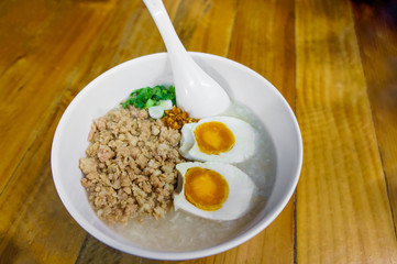 Minced pork congee with egg, fresh ginger and  coriander on white cup and spoon over the wood table. Second view.