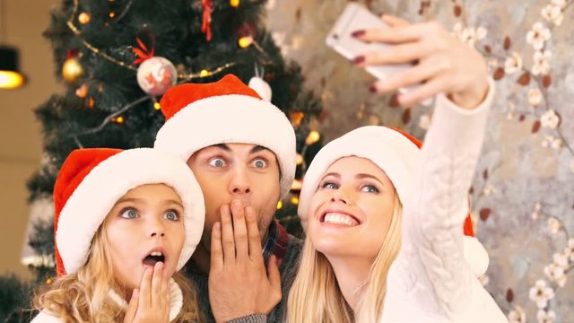 Playful young family in Santa's hat making selfie on smartphone near the Christmas tree