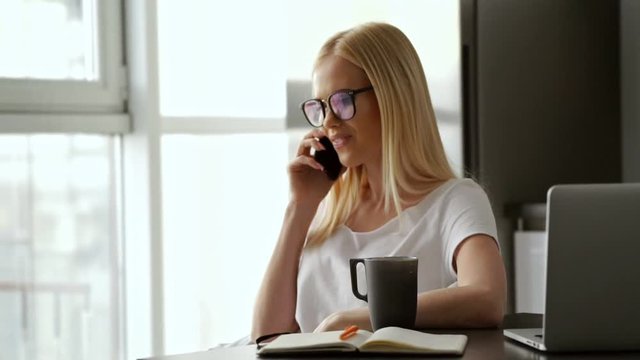 Cheerful blonde woman in eyeglasses sitting by the table indoors while talking by smartphone