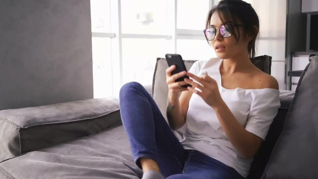Pretty asian woman in eyeglasses sitting on sofa at home while using smartphone