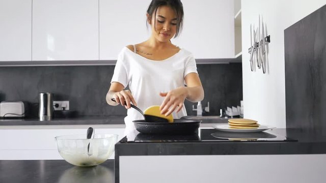 Playful asian woman standing by the stove on kitchen and cooking pancakes