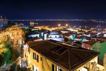 Illuminated old town of Nafplion in Greece with tiled roofs, small port, bourtzi castle, Palamidi fortress at night.